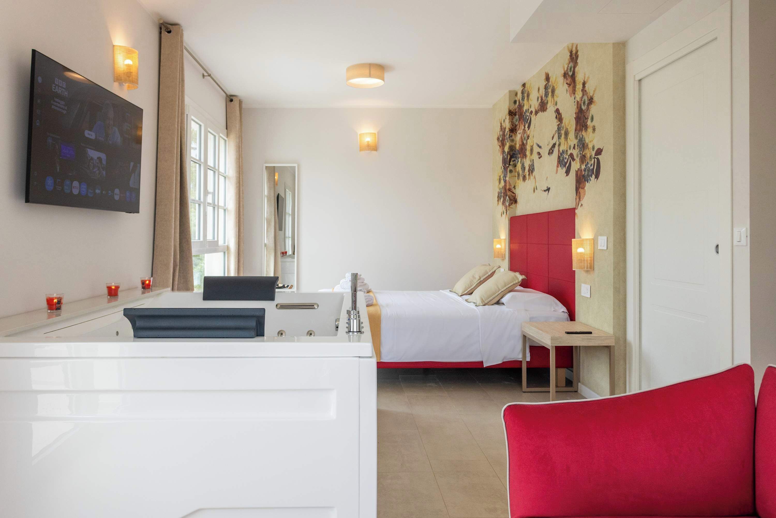 A hotel room with a bed, a desk, a mirror, a TV, and a red chair.