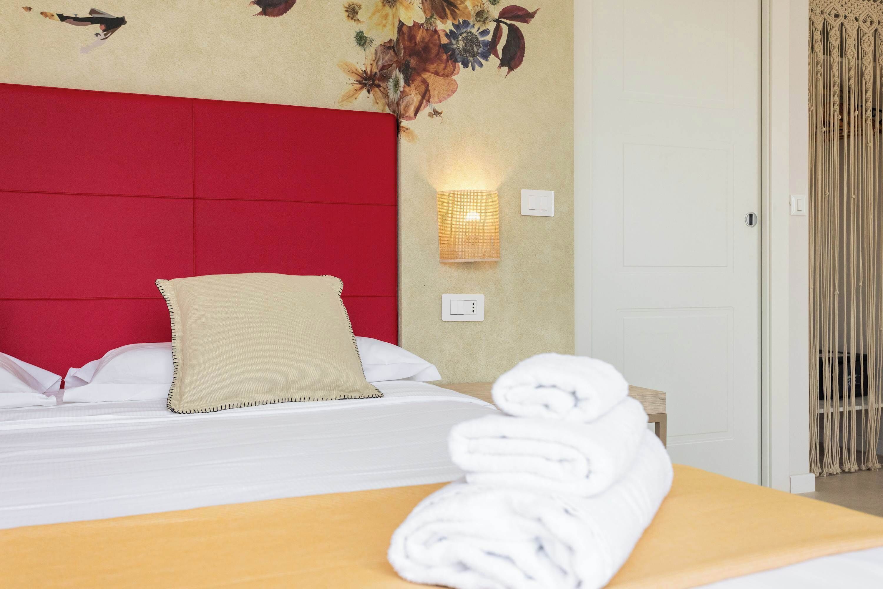 A hotel room with a red headboard, a wall-mounted light, a bed with white linens, and a rolled towel on a wooden table.