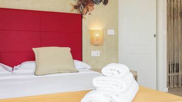 A hotel room with a red headboard, a wall-mounted light, a bed with white linens, and a rolled towel on a wooden table.