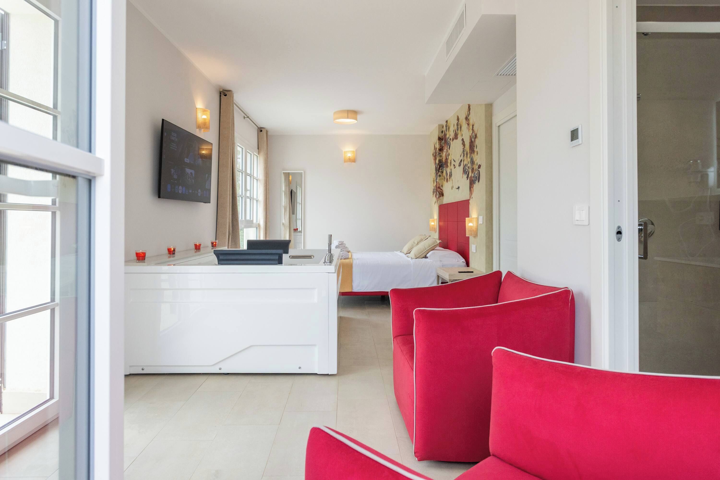 A modern hotel room with a bed, a desk, a red sofa, and a TV mounted on the wall.
