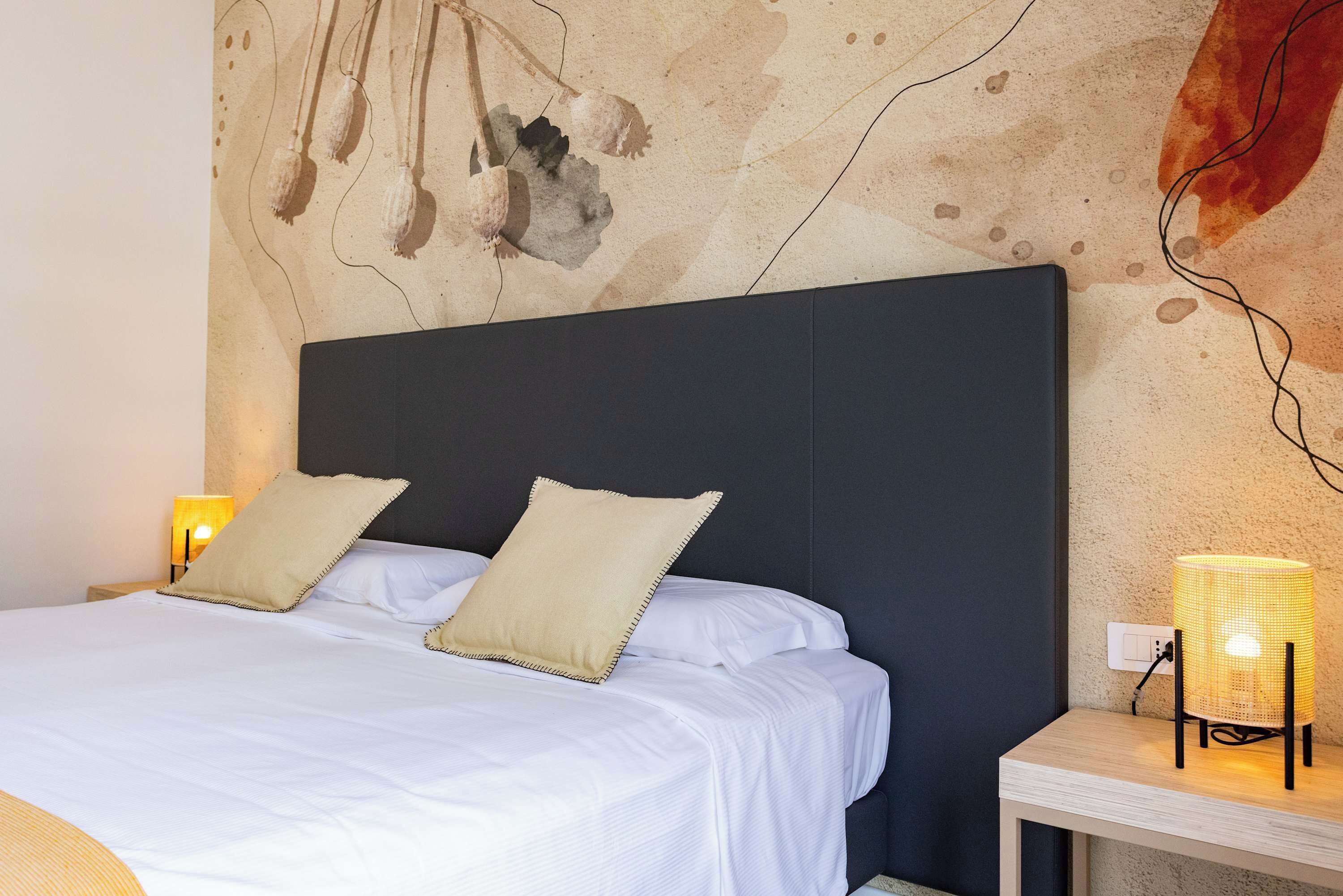 Una cama con sábanas blancas y dos almohadas color beige, un cabecero oscuro, una lámpara de noche y una pared con textura.