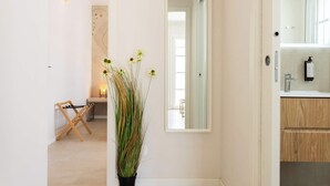 Un passadís amb una planta en test, un estendal de fusta i un mirall que reflecteix una habitació amb un rentanet i armaris de fusta.
