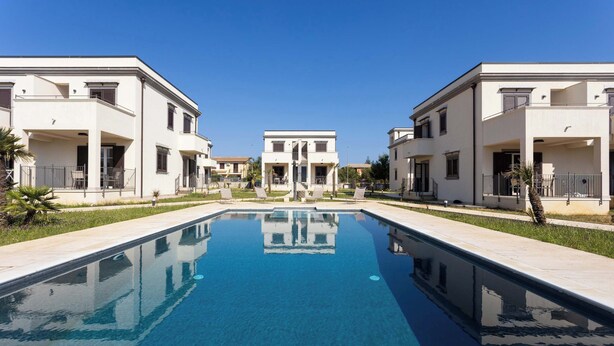 Una piscina amb el reflex d'edificis, envoltada d'una zona pavimentada.