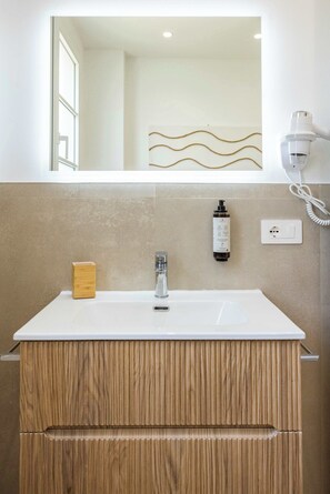 Ein Badezimmer mit einem weißen Waschbecken, einem Holz-Waschtisch, einem Spiegel, einem Haartrockner und einer Flasche Shampoo.