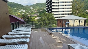 Un área de piscina en la azotea con sillones de descanso, piscina y vistas a edificios y colinas.