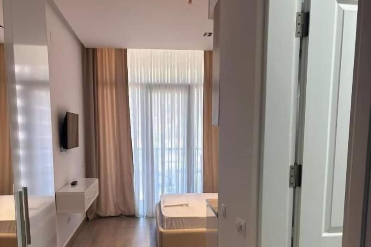 Una habitación de hotel moderna con cama, un pequeño escritorio, televisión y ventana con cortinas.
