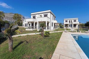 Une maison moderne à deux étages, dotée d’une piscine, entourée d’une pelouse bien entretenue et de palmiers.