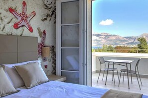 Una camera da letto con un letto, cuscini, un comodino, una lampada a parete e una porta-finestra scorrevole che dà su un balcone con tavolo e sedie.