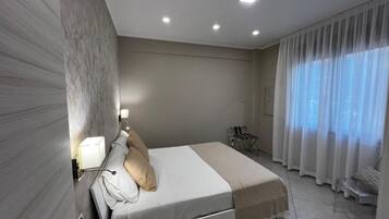 Un dormitorio moderno con cama, mesita de noche, lámpara y vistas al exterior a través de cortinas traslúcidas.