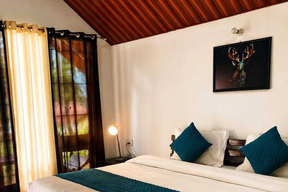 Um quarto com uma cama, mesa de cabeceira, abajur, obra de arte emoldurada e uma janela com cortinas.