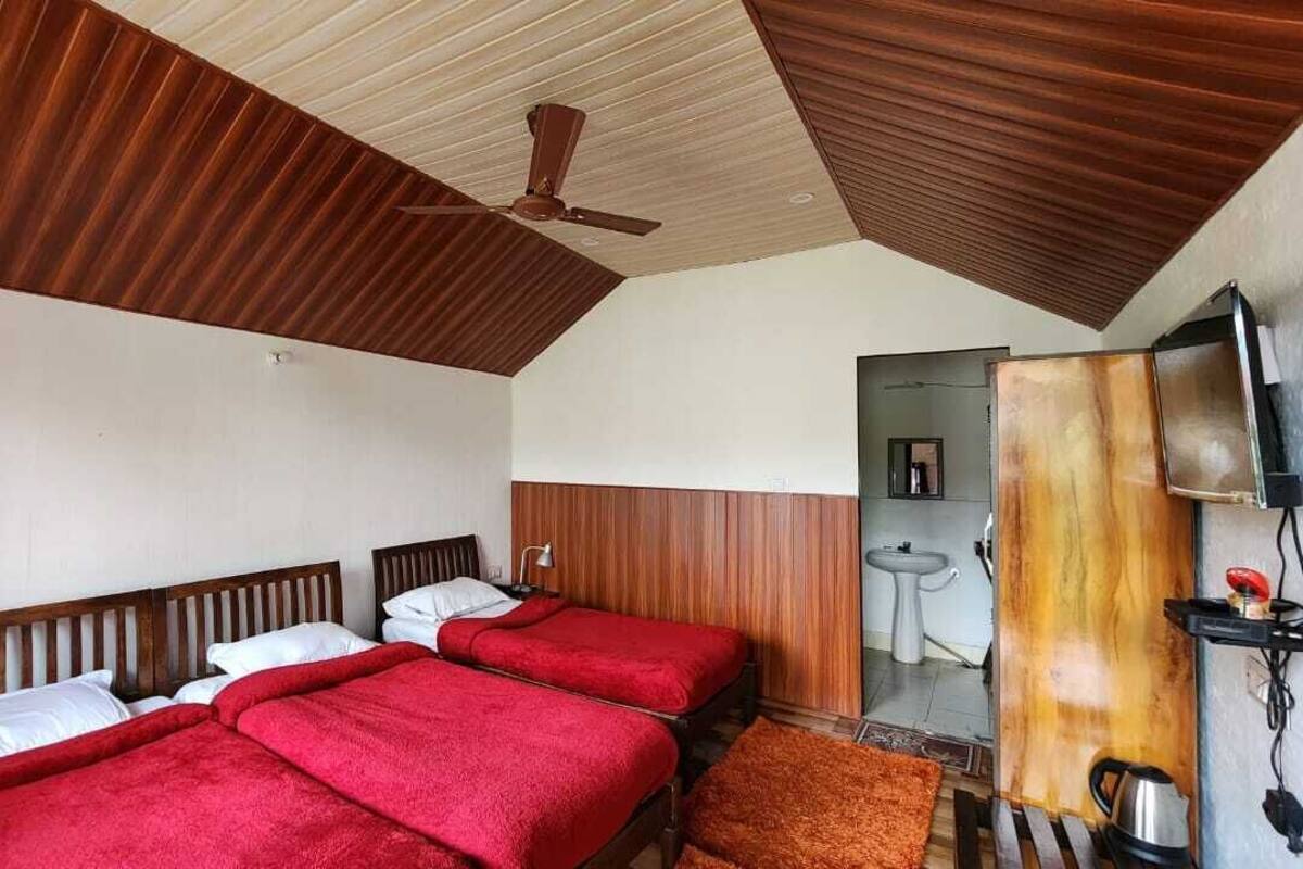 Quarto de hotel com duas camas, um ventilador de teto, teto de madeira, televisão e um banheiro pequeno.