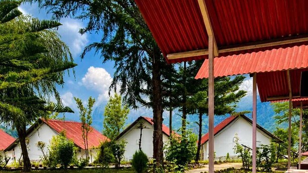 A covered outdoor seating area with wooden chairs and a checkered floor, surrounded by greenery and white buildings with red roofs. - Chardham Resort Guptakashi (Ukhimath)
