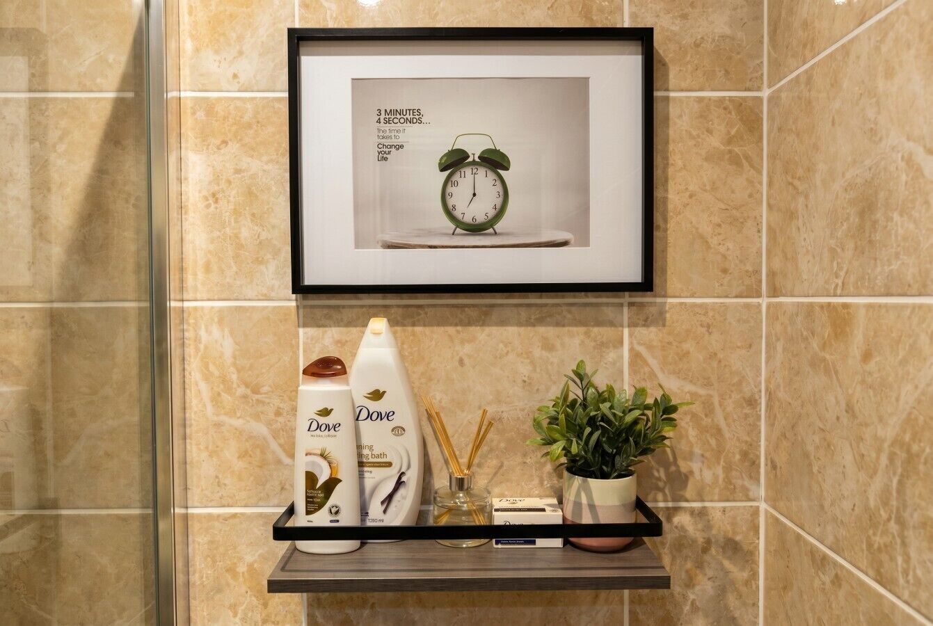 Une salle de bain avec une horloge encadrée, des produits de la marque Dove, une plante en pot et une cabine de douche en verre.