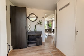 A hallway with a dark wooden door, a white wall with a geometric mirror, a small wooden console table with decorative items, and a dining area with chairs and a table in the background.