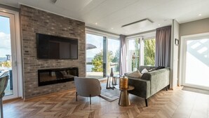 A modern living room with a fireplace, a sofa, a TV, and large windows offering a view of the outdoors.