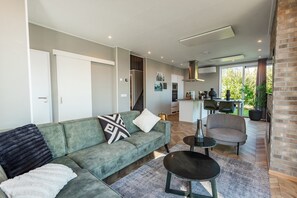 A modern living room with a sofa, a coffee table, and a chair. There is a kitchen area with a dining table and chairs visible through an open door.