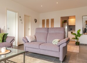 Ein modernes Wohnzimmer mit einer Couch, einem Glastisch und einer Küchenzeile im Hintergrund.