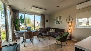 A modern living room with a sofa, chairs, a coffee table, and a dining area with a table and chairs. There is a large window with curtains, a floor lamp, and artwork on the walls. - De Wiedense Meren - Rietland Lodge | 4p (Wanneperveen)