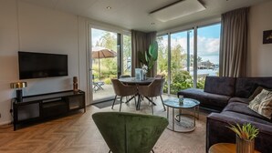 Una sala de estar moderna con una televisión de pantalla plana, un sofá, una mesa de comedor con sillas y vistas a una masa de agua a través de ventanas amplias.