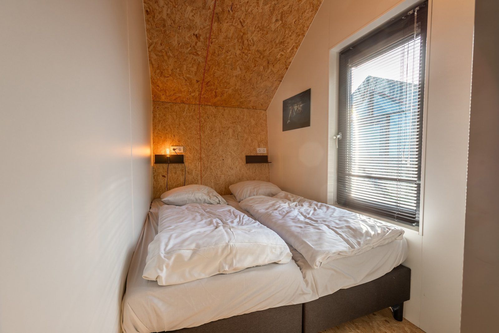 A bedroom with two beds, a wooden ceiling, a window with blinds, and a wall-mounted light.