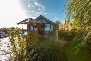 Une maison moderne à la façade en verre, située au bord d’un plan d’eau, avec des bateaux visibles au loin.