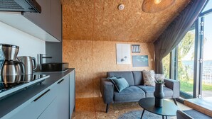 Et kompakt køkken med moderne hårde hvidevarer, en sofa med puder, et lille bord og udsigt til det fri gennem store vinduer.