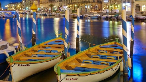 Una scena con vista sul mare, con barche ormeggiate a un molo, un albergo con insegne illuminate e una superficie d'acqua calma che riflette le luci.