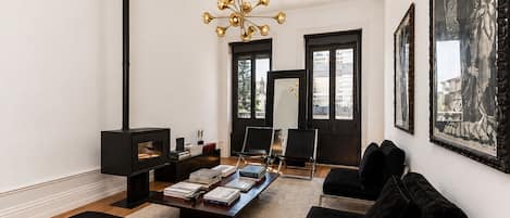 A modern living room with a fireplace, black sofas, a coffee table with books, a chandelier, and large framed artwork.