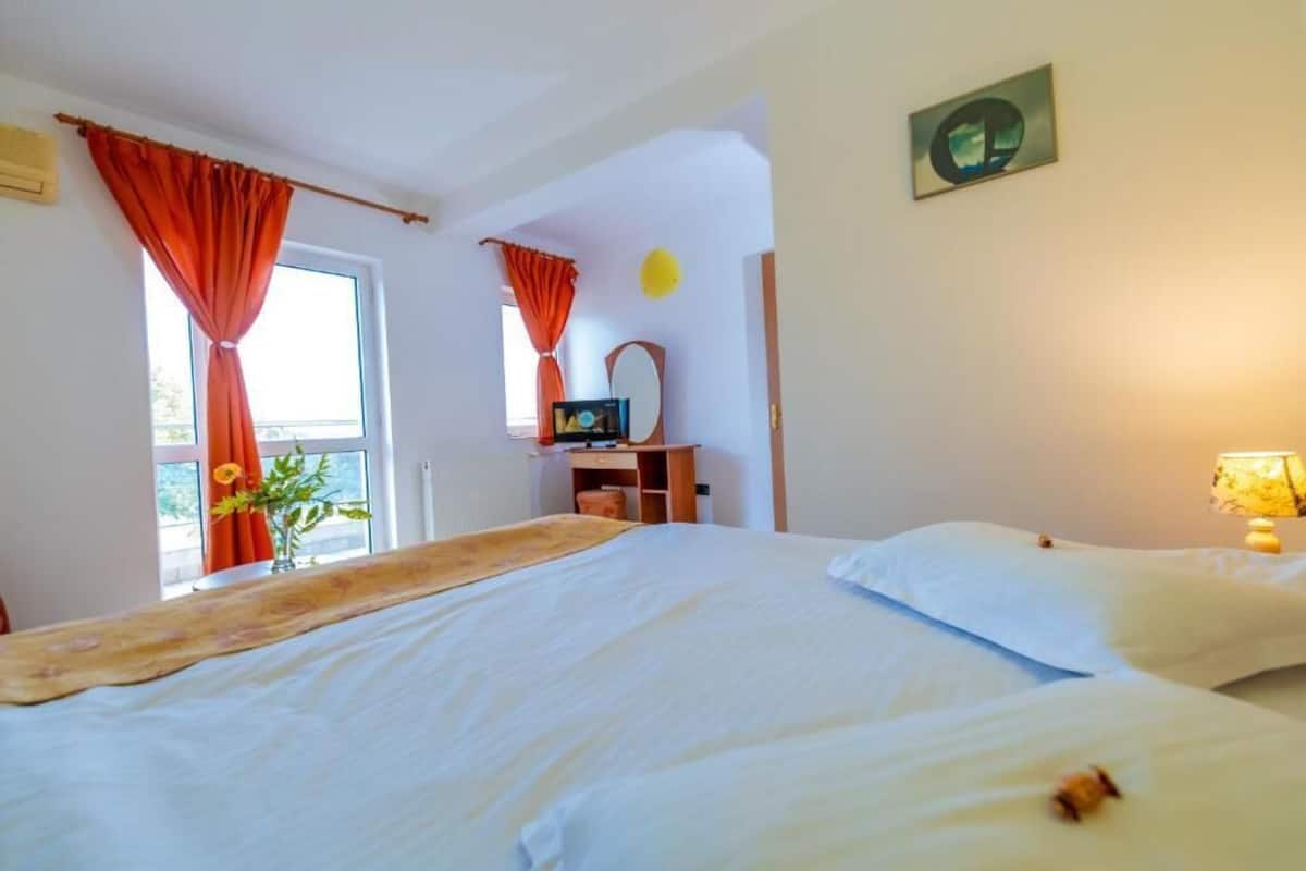 Ett hotellrum med en säng, ett skrivbord, en stol, ett fönster med gardiner, en växt och en tavla som hänger på väggen.