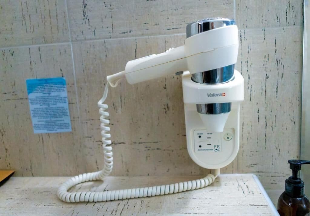 A white hair dryer with a cord, a power outlet, and a bottle of hand sanitizer on a wooden surface.