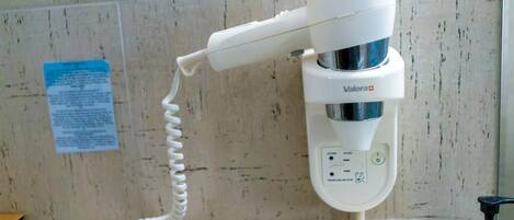 A white hair dryer with a cord, a power outlet, and a bottle of hand sanitizer on a wooden surface.