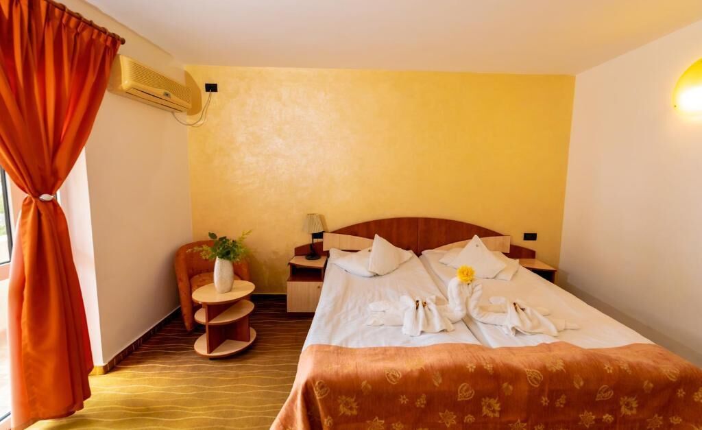 Una habitación de hotel con una cama, una mesita pequeña con una planta, una mesita de noche y un aire acondicionado instalado en la pared.