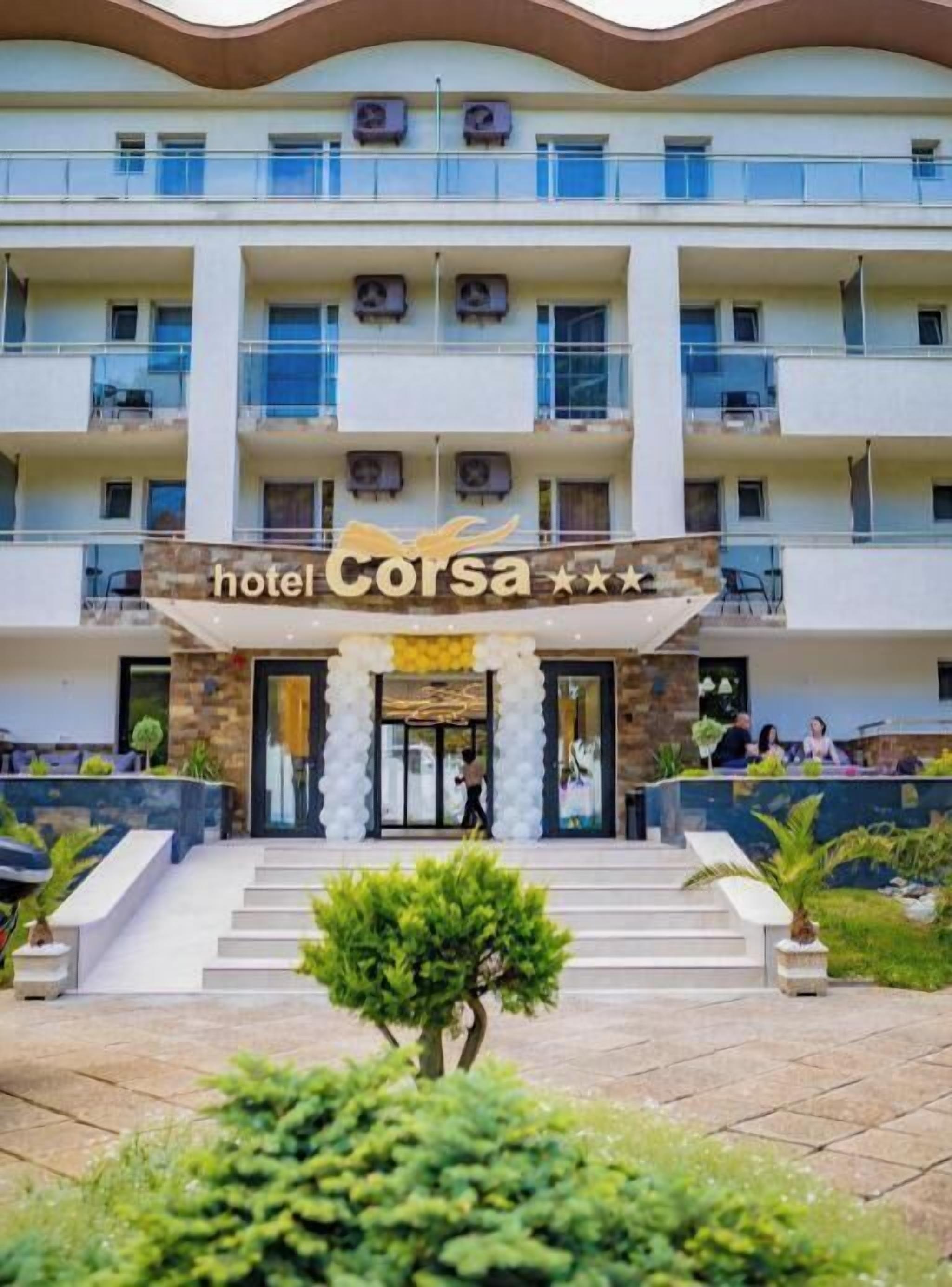 A multi-story hotel with a central entrance, balconies on each floor, and a landscaped front garden.
