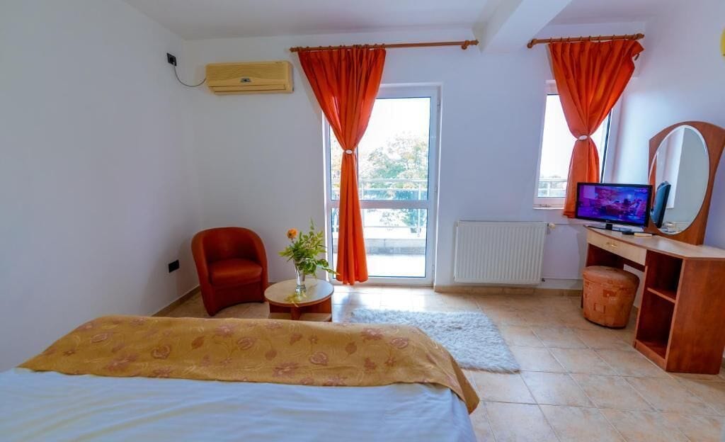 A bedroom with a bed, a chair, a desk with a TV, and a window with curtains.