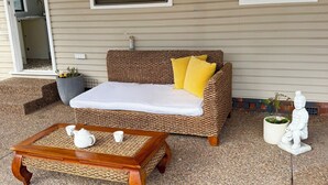 Eine Rattan-Couch mit weißem Kissen und gelbem Kopfkissen, ein Couchtisch aus Holz mit Teekannen und eine weiße Statue auf einer gefliesten Terrasse.