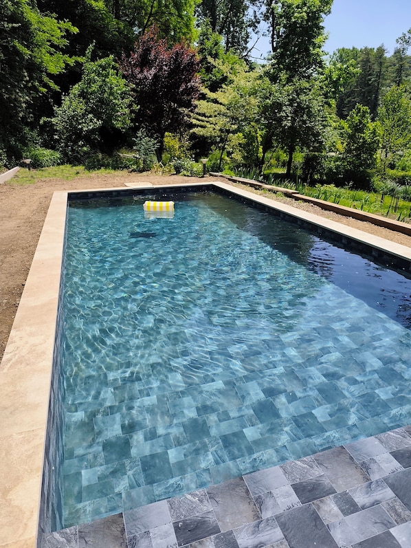 A rectangular swimming pool with clear water, surrounded by a tiled deck and lush greenery.