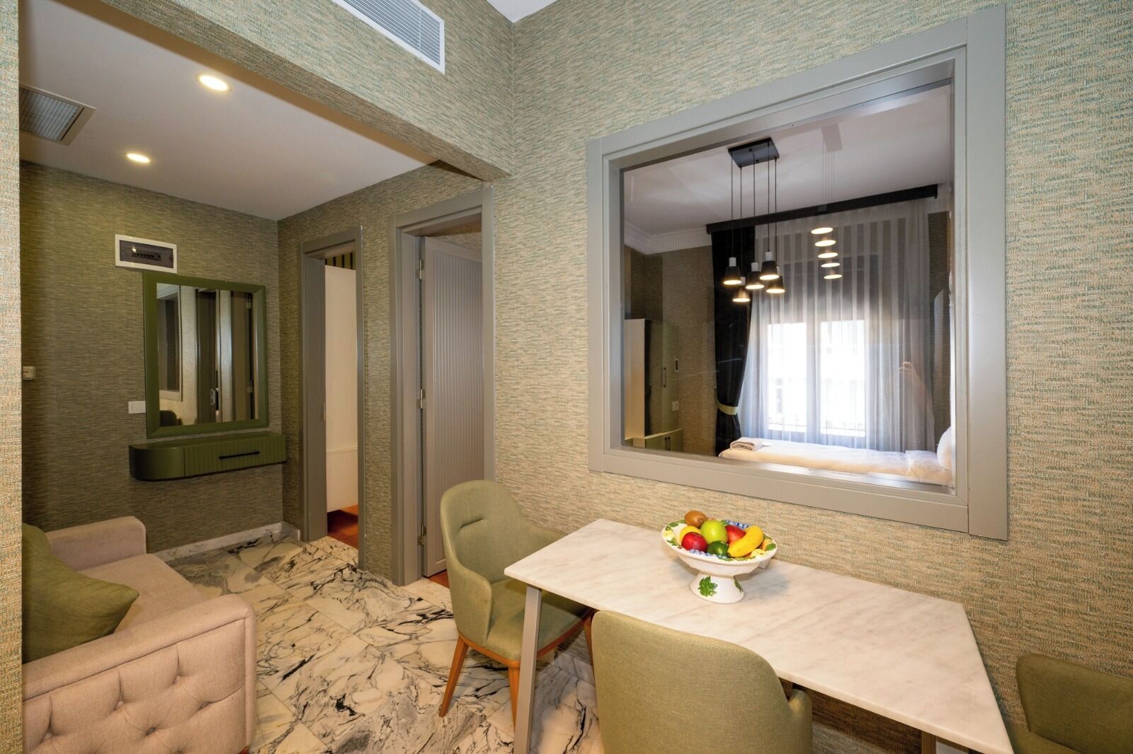 A hotel room with a marble floor, a dining table with a fruit bowl, a green armchair, a sofa, a large mirror, and a window with curtains.