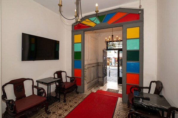 Une pièce avec un téléviseur, deux chaises en bois, une table, une moquette rouge et une porte colorée.
