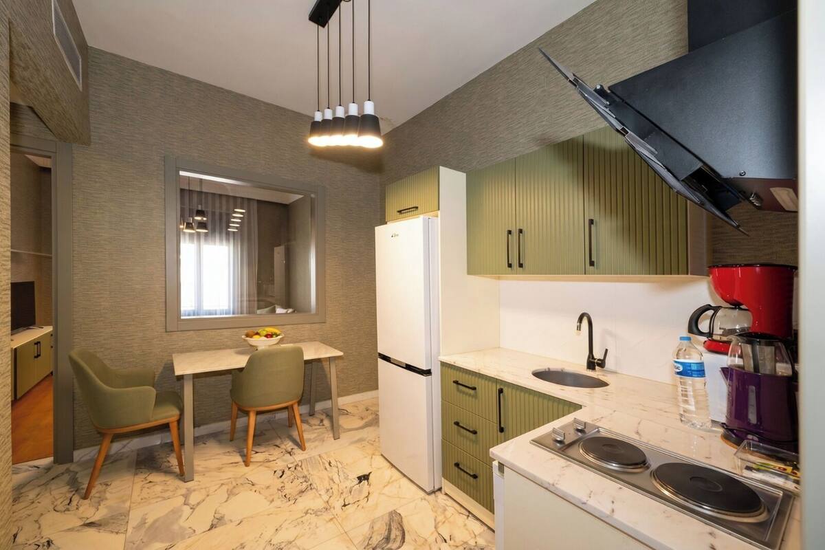 Cocina compacta con refrigerador, lavamanos y estufa, comedor con sillas y ventana con persianas.