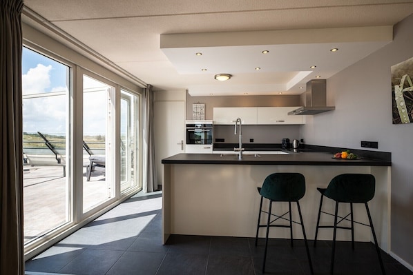 Una cocina moderna con una isla grande, dos taburetes de bar, un ventanal con vistas y un techo con iluminación empotrada.