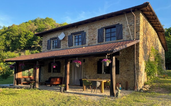 Une maison en pierre avec un toit en tuiles, une véranda avec des chaises et des jardinières suspendues.