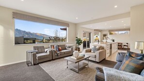 A spacious living area with a large window offering a mountain view, a kitchen with a dining area, and a separate seating space. - Alpine 75 Estate - Queenstown Hill 7 Bed (Queenstown)