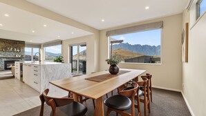A modern kitchen with a stone fireplace, a dining area with a wooden table and chairs, and a view of mountains through large windows. - Alpine 75 Estate - Queenstown Hill 7 Bed (Queenstown)