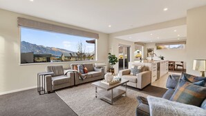 A spacious living area with a large window offering a mountain view, a kitchen with a dining area, and a separate seating space. - Alpine 75 Residence - Queenstown Hill 5 Bed (Queenstown)
