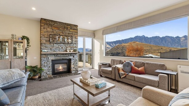A living room with a stone fireplace, a sofa, a coffee table, and a large window with a mountain view. - Alpine 75 Residence - Queenstown Hill 5 Bed (Queenstown)