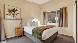 A bedroom with a bed, bedside table, lamp, and a window with curtains. - Alpine 75 Cottage - Queenstown Hill 2 Bed (Queenstown)
