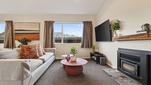 A living room with a sofa, a coffee table, a flat-screen TV, a fireplace, and a window with curtains. - Alpine 75 Cottage - Queenstown Hill 2 Bed (Queenstown)
