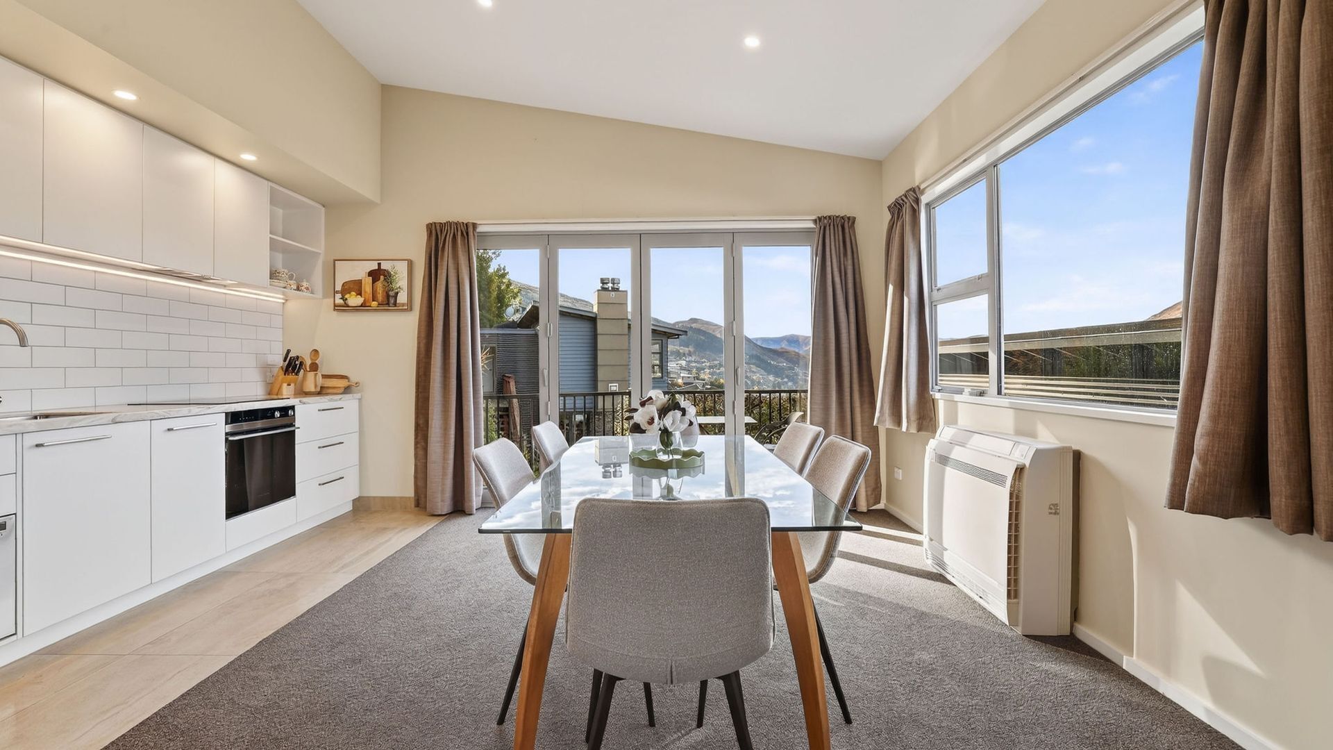 Une cuisine moderne avec des meubles blancs, une table à manger avec des chaises et une vue sur les montagnes grâce à de grandes fenêtres.