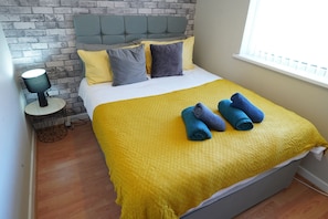 A neatly made bed with a yellow blanket and rolled towels, a grey headboard, and a brick-patterned wall. - Princetown Palace, Free Parking (Sunderland)