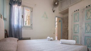 A bedroom with a large bed, a window with curtains, a fan, and a door with geometric designs. - Cozy and bright 80 square meters apartment, perfect for four people, located on the first floor of a condominium with a swimming pool (no elevator).The property is located in Liscia di Vacca, a short distance from Porto Cervo, reachable by car or public  (Liscia di Vacca)